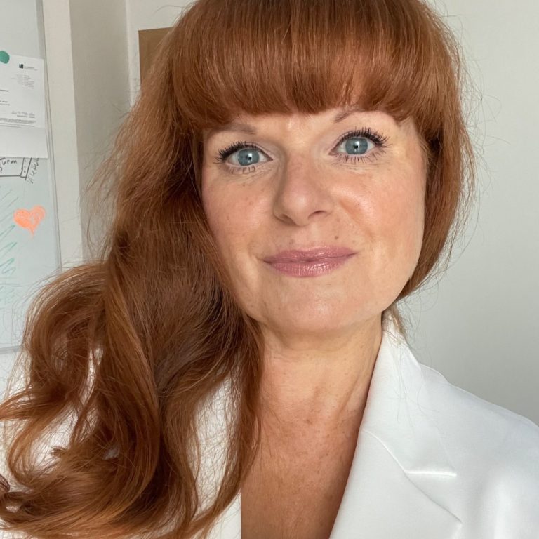 Beate Hinz mit langen, lockigen roten Haaren in einem weißen Blazer, lächelnd in die Kamera.