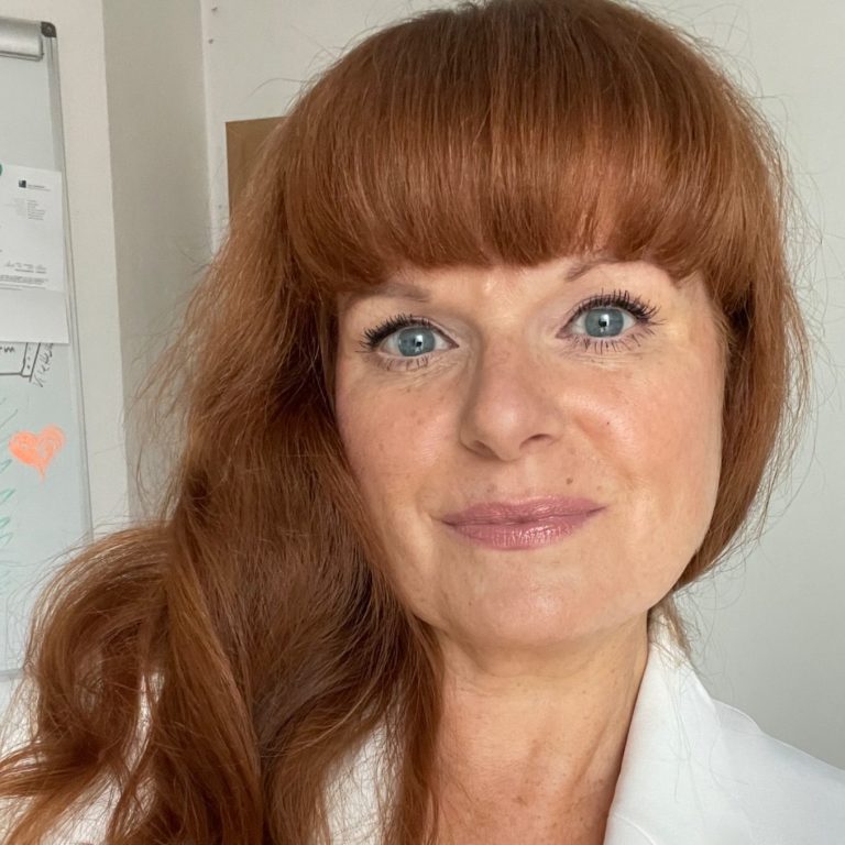 Beate Hinz Coaching und Training Beate Hinz mit langen, lockigen roten Haaren in einem weißen Blazer, lächelnd in die Kamera.
